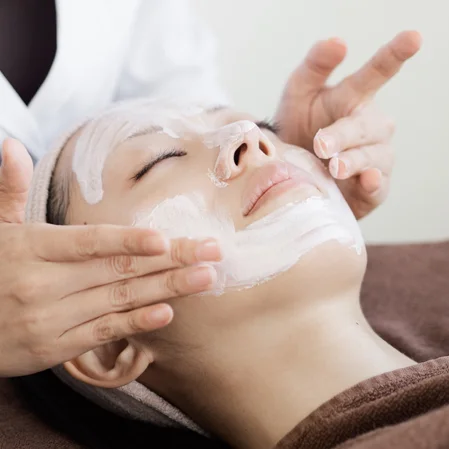 A woman lays on her back with her eyes closed. An esthetician applies a facial mask to her face