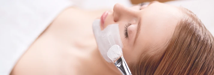 Massage Therapy Brentwood CA Esthetics A blonde haired woman lays on an esthetician table with her eyes closed. A brush applies a facial cream to her face.